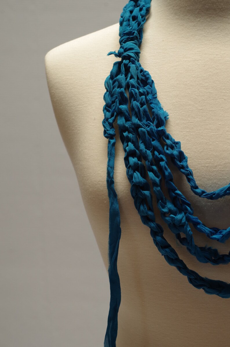 May include: A teal blue crocheted necklace with multiple strands of fabric. The necklace is displayed on a white mannequin.