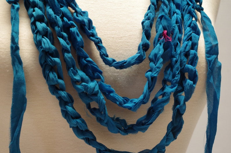 May include: A blue crocheted necklace made from fabric strips. The necklace is layered and has a braided design.