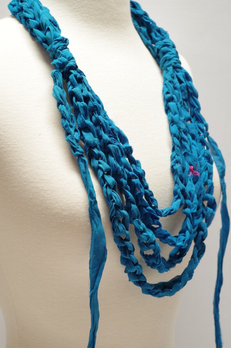 May include: A turquoise blue fabric necklace with multiple strands woven together. The necklace is displayed on a white mannequin.