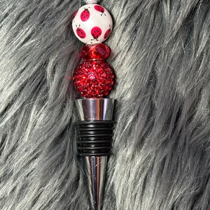 May include: A silver bottle stopper with a black rubber ring and three decorative beads. The top bead is white with red ladybugs, the middle bead is red, and the bottom bead is red with rhinestones.