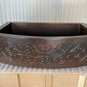 May include: A copper farmhouse sink with a hammered finish and a decorative scroll design. The sink is curved at the front and has a rectangular shape.