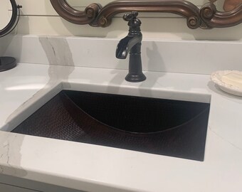 Puede incluir: Una encimera blanca con un lavabo de cobre marrón oscuro y un grifo negro. El lavabo está en un baño con un tocador blanco y un espejo grande y ornamentado.