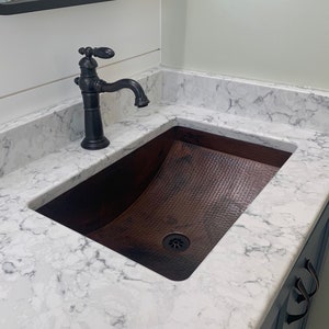 May include: A white countertop with a copper sink and a black faucet. The sink is hammered and has a slight curve to it. The faucet has a traditional design with a lever handle.