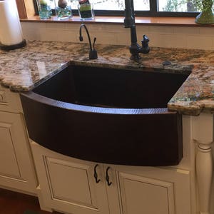 May include: A farmhouse style kitchen with white cabinets and a large copper farmhouse sink. The countertop is a multi-colored granite with flecks of brown, gold, and gray.