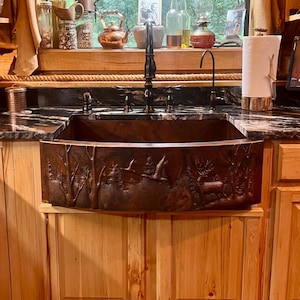 May include: A copper farmhouse sink with a detailed forest scene carved into the front. The sink is installed in a kitchen with light wood cabinets and a dark countertop. A black faucet is mounted above the sink.