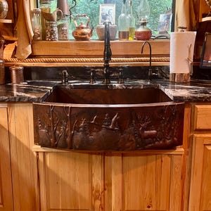 May include: A copper farmhouse sink with a detailed forest scene carved into the front. The sink is installed in a kitchen with light wood cabinets and a dark countertop. A window with a view of trees is above the sink.