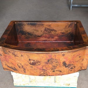 May include: A copper farmhouse sink with a hammered finish. The sink has a curved front edge and a rectangular shape.