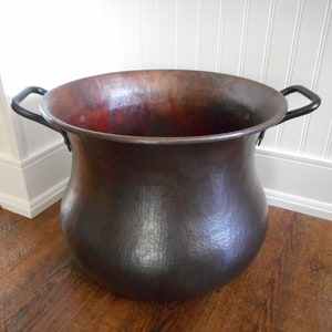 May include: A large, hammered copper planter with two black iron handles. The planter has a wide, rounded base and a narrow top.