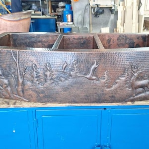 May include: A hammered copper kitchen sink with a rectangular shape and three compartments. The front of the sink features an embossed nature scene with deer, trees, and flying birds. The sink is displayed on a blue cabinet.