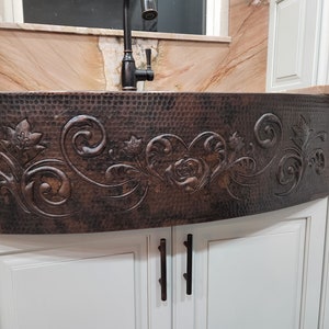 May include: A copper farmhouse sink with a hammered finish and an ornate floral design. The sink is installed in a white kitchen cabinet with black hardware.