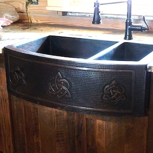May include: A double copper farmhouse sink with a hammered finish and Celtic knot designs. The sink is installed in a wooden countertop.