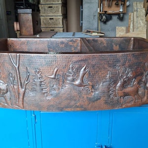 May include: A large, copper farmhouse sink with a hammered finish and an embossed design of trees and deer. The sink is double-bowl and has a curved front edge.