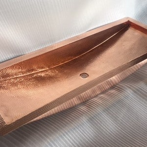 May include: A rectangular copper sink with a hammered finish. The sink has a curved shape and a drain hole in the center.
