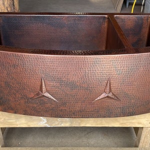 May include: A copper farmhouse sink with a hammered texture and a double basin design. The front of the sink features three star-shaped cutouts. The sink is supported by a wooden frame.