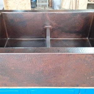 Puede incluir: Un fregadero de cocina de cobre de doble seno con una textura martillada. El fregadero rectangular tiene un divisor central y un borde ancho. El cobre tiene un tono marrón rojizo.