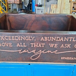 May include: A copper farmhouse sink with a hammered finish. The sink has an inscription that reads "Exceedingly, Abundantly Above All That We Ask Or Imagine Ephesians 3:20".