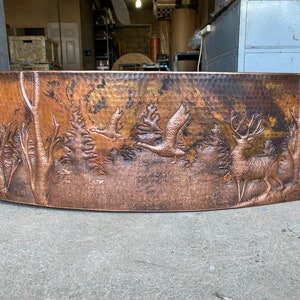 May include: A copper farmhouse sink with a hammered finish and an embossed design of trees, birds, and a deer. The sink is curved and has a rustic look.