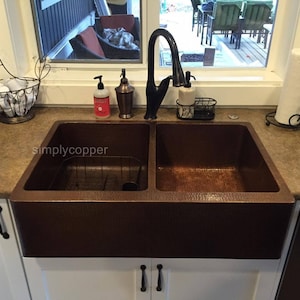 Puede incluir: Un fregadero de granja de cobre de doble cubeta con acabado martillado. El fregadero está instalado en una encimera de cocina con gabinetes blancos y una encimera marrón. El fregadero tiene un grifo simple con un caño de cuello de cisne.