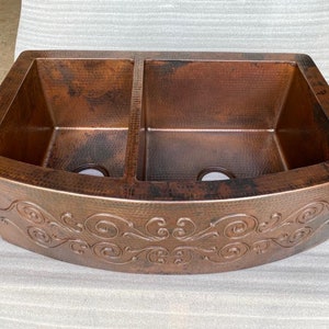 May include: A copper double-basin farmhouse sink with a hammered texture and decorative scrollwork along the apron. The sink has a rich, warm brown color with darker accents, and two drain holes are visible.