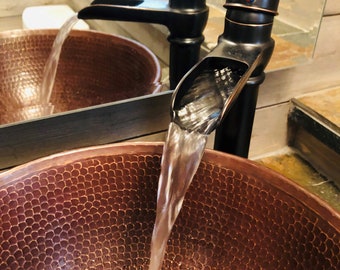 May include: A close-up of a copper sink with a waterfall faucet. The faucet is black and has a decorative design. Water is flowing from the faucet into the sink.