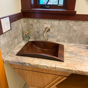 May include: A copper sink with a hammered finish sits on a countertop with a brown and white marbled pattern. The sink is mounted on a wooden cabinet with a geometric design.