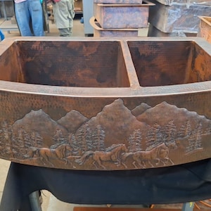 May include: A double-basin copper kitchen sink with a hammered finish and a mountain and horses design. The sink is made of solid copper and has a rustic look.