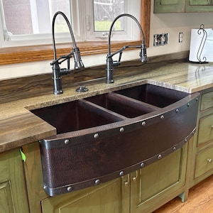 May include: A copper farmhouse sink with a three-basin design, accented with rivets along the curved front. Two black gooseneck faucets are installed above the sink, set against a granite countertop and green cabinetry. The kitchen features a window and a paper towel holder.