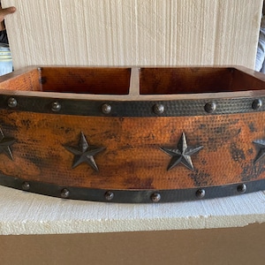 May include: A copper farmhouse sink with two basins and a hammered finish. The sink has a black iron rim with decorative star accents and rivets.
