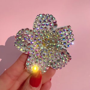 Puede incluir: Un broche de color blanco con forma de flor con diamantes de imitaci&oacute;n iridiscentes. El broche est&aacute; hecho de un material blanco y est&aacute; cubierto de peque&ntilde;os diamantes de imitaci&oacute;n brillantes.
