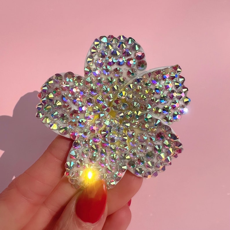 Puede incluir: Un broche de color blanco con forma de flor con diamantes de imitaci&oacute;n iridiscentes. El broche est&aacute; hecho de un material blanco y est&aacute; cubierto de peque&ntilde;os diamantes de imitaci&oacute;n brillantes.