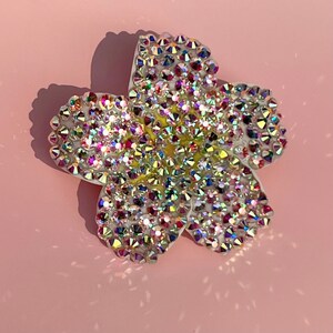 Puede incluir: Un broche de color blanco con forma de flor con pedrer&iacute;a iridiscente. La pedrer&iacute;a est&aacute; dispuesta en un patr&oacute;n que se asemeja a una flor.