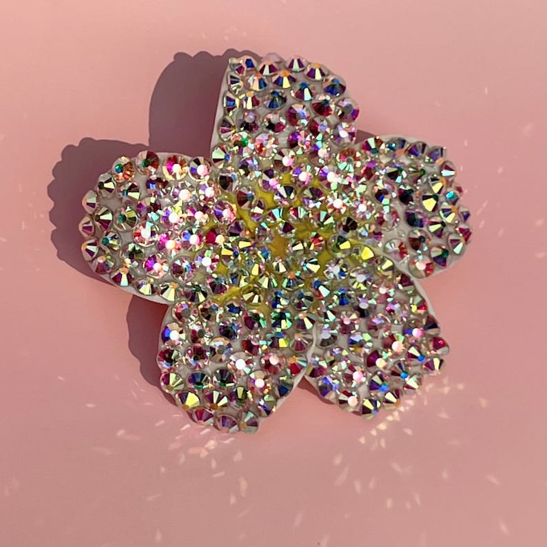Puede incluir: Un broche de color blanco con forma de flor con pedrer&iacute;a iridiscente. La pedrer&iacute;a est&aacute; dispuesta en un patr&oacute;n que se asemeja a una flor.