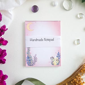 May include: A handmade notepad with a pink and white gradient background, featuring a crescent moon and floral illustrations. The notepad is wrapped with a white band that reads "Handmade Notepad". Decorative elements include purple flowers, glass beads, and a green ribbon.