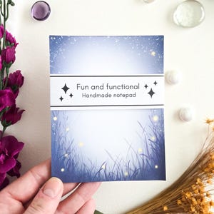 May include: A handmade notepad with a blue and white design, featuring a starry sky and tall grass illustration. The notepad has the text "Fun and functional Handmade notepad" on a white band. The notepad is held by a hand.