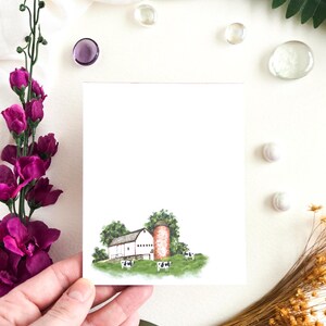 May include: A watercolor illustration of a farm scene featuring a white barn, a red silo, and cows grazing on a green hill. The artwork is on a white card, held by a hand, with purple flowers and decorative elements in the background.