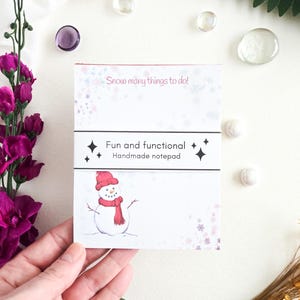 May include: A handmade notepad with a snowman illustration wearing a red hat and scarf. The notepad has the text "Snow many things to do!" and "Fun and functional". The notepad is white with pink and purple accents.