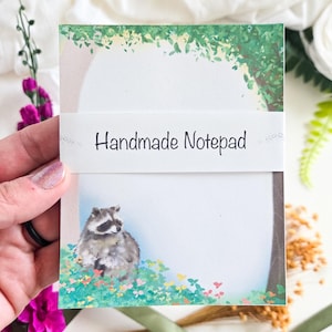 May include: A handmade notepad featuring a watercolor illustration of a raccoon in a field of flowers. The notepad has a white background with a green and brown tree design at the top. The words "Handmade Notepad" are printed on a white band.