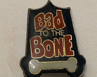 May include: A black enamel pin with a gold outline. The pin features a white bone with a red and gold "Bad to the Bone" text.