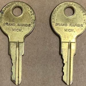 Puede incluir: Dos llaves doradas con el texto "AMERICAN METAL HDWE. CO. GRAND RAPIDS, MICH." en la parte superior de cada llave.