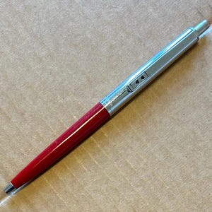 May include: A red and silver ballpoint pen. The pen features a red barrel and a silver metal top and tip. The pen is positioned on a brown surface.