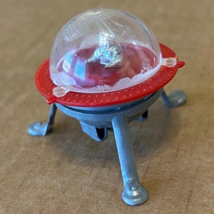 Op de afbeelding: Een kleine speelgoed ruimteschip met een doorzichtige koepel en een rode cirkelvormige basis. Het ruimteschip heeft drie grijze poten en een kleine zilveren figuur binnenin. De basis heeft een rode ring met kleine gaatjes rond de rand.