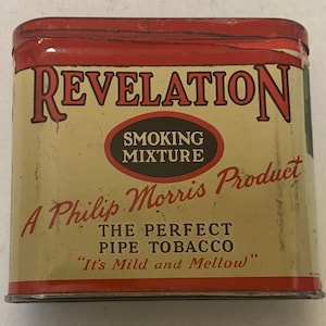 May include: Vintage Revelation smoking mixture tin. The rectangular tin has a red top and a cream-colored body with red lettering. The text includes "REVELATION", "SMOKING MIXTURE", and "A Philip Morris Product". The tin also states "THE PERFECT PIPE TOBACCO".