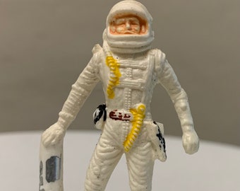 Könnte beinhalten: Eine weiße Plastik-Astronautenfigur mit einem gelben und braunen Gürtel, einem weißen Helm mit rotem Gesicht und einem silbernen Werkzeug in der Hand.