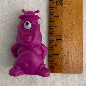 May include: A purple plastic toy alien with one large eye, a small mouth, and two antennas on its head. The toy is about 2 inches tall.