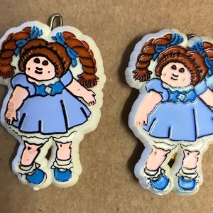 May include: Two plastic doll ornaments with brown hair, blue dresses, and white shoes. The dolls are smiling and have a blue bow on their heads.