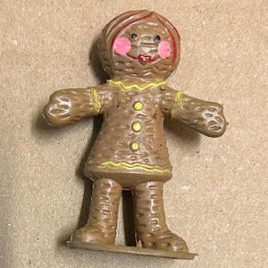 May include: A brown gingerbread person figurine with yellow icing details and pink cheeks. The figurine has a red hair bow and is standing on a brown base.