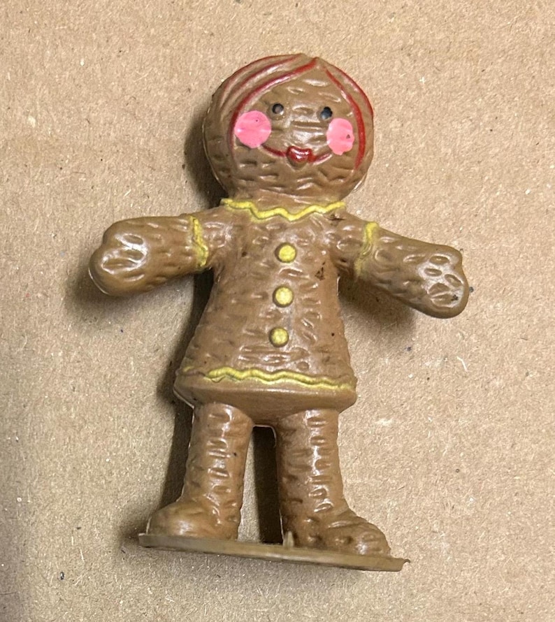 May include: A brown gingerbread person figurine with yellow icing details and pink cheeks. The figurine has a red hair bow and is standing on a brown base.