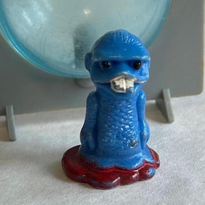 May include: A small, blue figurine with a creature-like design. The figure has a textured body, a small head with black eyes, and a white-toothed mouth. It stands on a red base. The background is a light blue and gray.