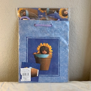 Op de afbeelding: Een blauwe cadeauzak met een zonnebloemdessin en de tekst "Anne Geddes" erop gedrukt. De tas heeft een afbeelding van een baby die een zonnebloemkostuum draagt en in een bloempot zit.