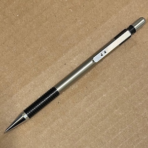 May include: A silver mechanical pencil with a black textured grip and a conical tip. The pencil has a metallic finish and a black band near the eraser. The pencil is lying on a brown surface.